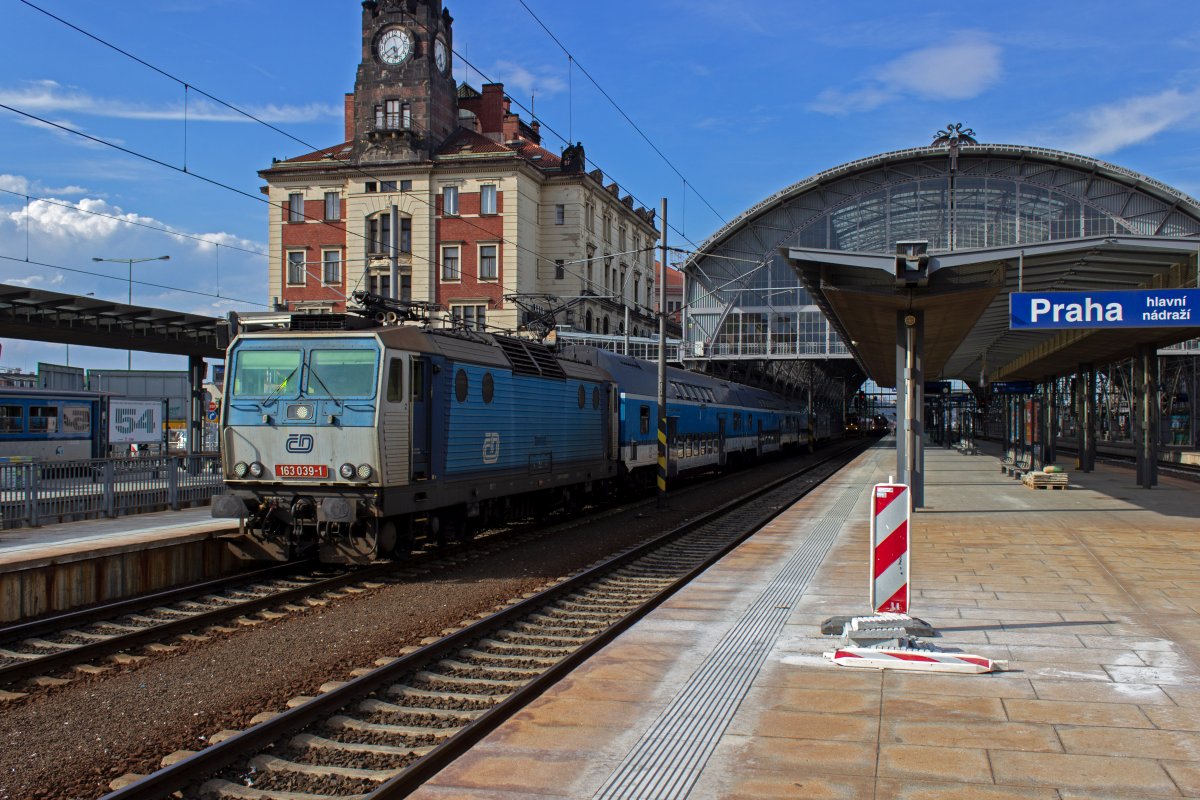 163 039 startet mit einem Verst�rkungszug der Linie S7 nach Řevnice am Hausbahnsteig des Prager Hauptbahnhofs. Aufgrund der eigenwilligen Nummerierung von Bahnsteiggleisen in Tschechien handelt es sich dabei um Gleis 9, rechts davon die Gleise 7 und 1. Zur Vereinfachung f�r die Fahrg�ste wird �blicherweise nur der Bahnsteig an dem der Zug abf�hrt angezeigt. Die Gleisnummern folgen aber durchaus einer Regel: Die durchgehenden Hauptgleise hei�en 1 und 2, von dort ausgehend wird auf je einer Seite mit geraden, bzw. ungeraden Nummern weitergez�hlt, wobei Abstell- und Betriebsgleise mitgez�hlt werden.