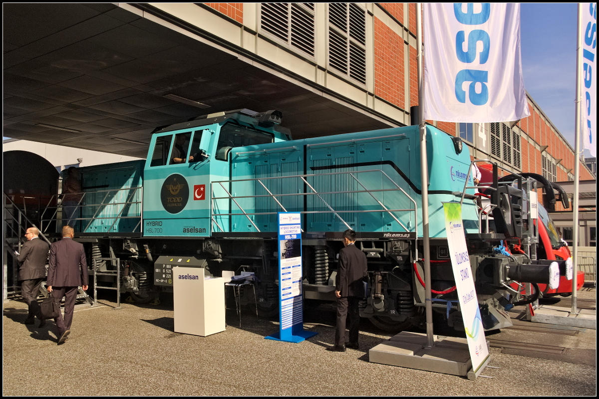 Eng ging es zu auf der Freifläche auf der InnoTrans 2018 am 18.09.2018. So ergaben sich wenig Lücken um die ausgestellten Loks und Wagen zu fotografieren. Die vom türkischen Konsortium Aselsan und Tülomsas gebaute HSL-700 fährt sowohl mit Diesel (300 kW) als auch mit Lithium-Ionen-Batterie (400 kW). Eingesetzt wird sie bei der TCDD als Rangierlokomotive und wird von den Herstellern auch als solche angeboten. Mit einem Gewicht von 68 t kommt sie auf Vmax 80 km/h. Beide Antriebssysteme können getrennt, aber auch zusammen betrieben werden.