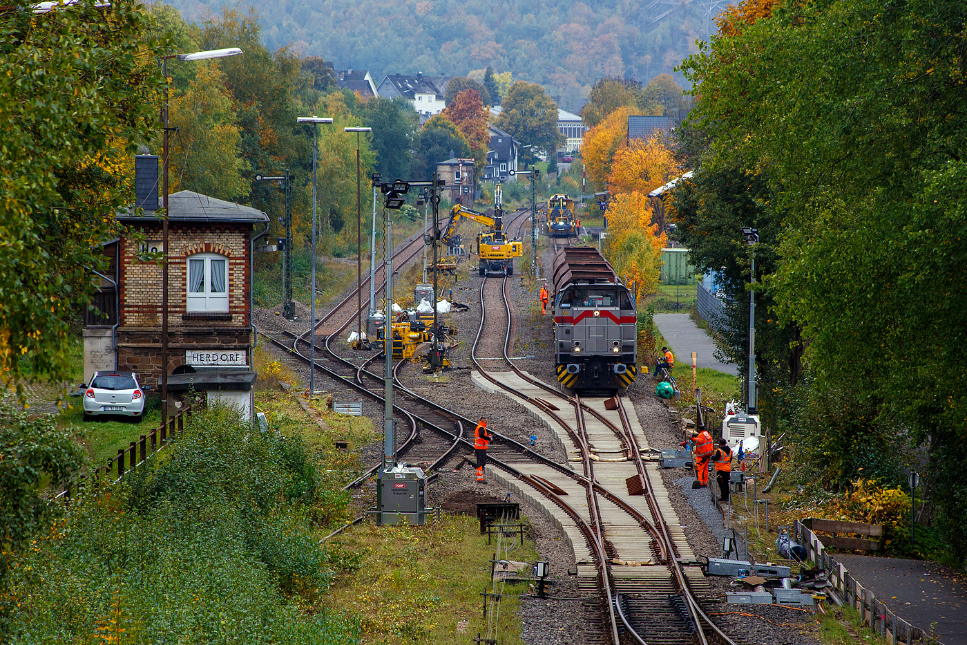 Blick auf den Bahnhof Herdorf und die Baustelle in Blickrichtung Betzdorf am 12 Oktober 2025. Die Weichen 25 und 26 sind beide eingebaut, müssen noch eingeschottert werden.

So steht die 277 809-0  Elmi“ (92 80 1277 809-0 D-KAF), eine Vossloh MaK G 1700 BB der KAF - Falkenhahn Bau AG (Kreuztal), steht mit einem Schotterzug bereit. Hinten beim Bahnhof haben die Schnellschotterplaniermaschine SSP 110 SW, Schweres Nebenfahrzeug Nr. 99 80 9425 068-0 D-DGU und die Universalstopfmaschine UNIMAT 09-475/4S, Schweres Nebenfahrzeug Nr. D-DGU 99 80 9424 001-2, den Bahnhof erreicht müssen aber nach das Einschottern abwarten. Beide Plasser & Theurer Maschinen gehören der Deutsche Gleisbau Union GmbH & Co. KG (DGU), Koblenz

Hier sieht man außerdem gut das Herdorf immer noch den Luxus von 2 Stellwerken besitzt. Vorne links das Weichenwärter Stellwerk Herdorf Ost (Ho) und hinten das Stellwerk Herdorf Fahrdienstleiter (Hf). Beide Stellwerke waren sogar hier bei der Baustelle ganztäglich besetzt.

Ich denke das bei den heutigen Verhältnissen im Bahnhof Herdorf, mit drei aktiven Gleisen und einer Anschlussstelle zum Rangierbahnhof der KSW Kreisbahn Siegen-Wittgenstein (Betriebsstätte FGE - Freien Grunder Eisenbahn) nicht mehr zeitgemäß ist. Es gibt mit Sicherheit heute Möglichkeiten die Aufgaben von einem oder dem anderen Stellwerk fernzusteuern. Früher mit bis zu 30 Gleisen und 3 Anschlussstellen musste es so sein, aber im heutigen Zustand