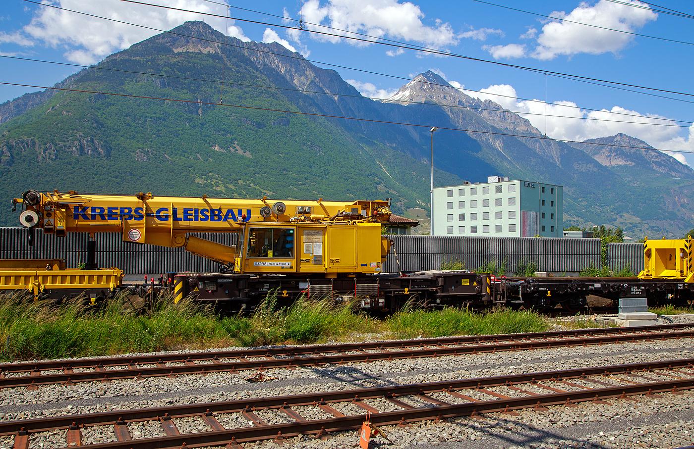 Der 130 t Eisenbahnkran D-KREBS 99 80 9419 008-4, ex 97 82 54 501 17-1, ein GOTTWALD GS 130.06 TT Gleisbauschienenkran der Krebs GmbH & Co. KG, Bingen am Rhein (D) / Bern (CH) ist am 26.05. 2023 beim Bahnhof Martigny, in Transportstellung mit dem Gegenlastwagen 33 88 3936 639-4 B-Krebs der Gattung Rels und dem Schutzwagen auslegerseitig 99 80 9383 003-7 D-Krebs der Gattung Rgs, abgestellt.

Der Kran wurde 2001von GOTTWALD (Mannesmann Dematic) in Dsseldorf unter der Fabriknummer 846 375gebaut.

Der GS 130.06 TT ist eine Weiterentwicklung des GS 80.08 T. Folgende Verbesserungen wurden vorgenommen, die den Einsatzbereich des Kranes nochmals erweitern:
• Der Einsatz eines dreiteiligen Auslegers (ein Grundschuss und zwei Teleskopschsse) ermglicht eine wesentliche Erweiterung des Arbeitsbereiches. Durch die krzere Bauform des Auslegers beginnt der verfgbare Bereich horizontal bereits 3,0 m vor Puffer mit einer Tragfhigkeit von 98,0 t (fr
• eistehend 78,0 t). Damit ist er der einzige am Markt verfgbare Gleisbaudrehkran, der diese, gerade fr den Brckenbau, wesentliche Fhigkeit besitzt.
• Erweiterung des Ausladungsbereiches vor Puffer um 2,0 m auf 15,5 m max. Ausladung.
• Erhhung der Tragfhigkeit bei max. Ausladung vor Puffer von 15,5 m auf 36 t.
• Fr Schwerlastflle ist der Kran mit einem zustzlichen Anschlagpunkt versehen, wodurch es jetzt mglich ist, Lasten von 100 t im 7 m Radius, bis zu einem Schwenkwinkel von 30  aufzunehmen und zu bewegen.
• Verwendung geschlossener und damit wartungsarmer Fahrgetriebe. Selbstfahrgeschwindigkeit bis max. 25 km/h (zugel. 20 km/h) sind damit mglich.
• Der Kran verfgt ber die Mglichkeit mit nur einer Absttzung zu arbeiten, entsprechende Traglasttabellen sind in die Lastmomentberwachung eingearbeitet. Die Absttzungen werden von der Fahrerkabine aus bettigt und berwacht.

Der GS 130.06 TT verfgt selbstverstndlich ber alle weiteren Ausstattungen, die bereits vom GS 80.08 T bekannt sind und diese Krane gerade fr den Gleis- und Brckenbau so vielseitig und sicher einsetzbar machen, sind wie u.a. den automatischen Gleisberhhungsausgleich, jetzt aber bis 180 mm Gleisberhhung, die elektronische berwachung und Steuerung des Lastmomentes unter Einbeziehung der Gegengewichts- und Absttzposition u.v.a.m. Die berhhungsausgleich-Einrichtung stellt den Kran immer waagerecht. Somit entfllt die Reduzierung der frei stehenden Tragfhigkeiten.

Fr diesen Krane sind entsprechend Einsatzzweck Traversen unterschiedlicher Tragfhigkeit und Abmessungen verfgbar.
Dieser Kran ist in die Streckenklasse CE/CM4 eingestuft, das bedeutet, er kann in Regelzgen bis 120 km/h eingestellt werden. Bei berstellfahrten muss keine BZA beantragt werden (Schwerlast). Dieser Kran hat durch Ablegen vom Gegengewicht (am Gegengewichtwagen) eine Achslast von 13,5 t.

In Transportstellung wird der Kran mit einem vorangestellten 4-achsiger Schutzwagen auslegerseitig, beladen u.a. mit den zugeh. Traversen, und hier in der Transportstellung wird der Kranausleger hydraulisch auf dem Schutzwagen abgesttzt, sowie dem vierachsigen Gegenlastwagen (ein Drehgestell-Flachwagen), auf dem Wagen liegen die Gegengewichte (Gegenlast) zum Kran. Durch die Absttzung wird die Achslast und folglich die Streckenlast (Meterlast) vom Kran reduziert. Zudem kann eine Lastablegung (Gegengewicht) auf Gegengewichtswagen im Kranbetrieb fr eine Achslastreduzierung erfolgen.

TECHNISCHE DATEN:
Spurweite: 1.435 mm (Normalspur)
Anzahl der Achsen: 8 (in 4 Drehgestelle bzw. 2 Doppeldrehgestelle)
Unterwagenlnge ber Puffer: 13.000 mm
Breite in Transportstellung: 3.100 mm
Hhe in Transportstellung: 4.250 mm
Drehzapfenabstand: 8.000 mm
Drehzapfenabstand im Doppeldrehgestelle: 2.300 mm
Achsabstand im Einzel-Drehgestell: 1.100 mm
Laufkreisdurchmesser der Rder: 730 mm (neu) / 680 mm (abgenutzt)
Gesamtgewicht  in Transportstellung:108 t (Gegengewicht abgelegt)
Grter Achsdruck in Transportstellung: 13,5 t
Schwenkwinkel des Oberwagens: 30 
Max. hintere seitlich Ausladung: 2.000 mm
Maximale Ausladung vor Puffer : 18 m
Kleinster befahrbarer Bogenradius: 90 m
Hchstgeschwindigkeit: 20 km/h (eigen / BB 25 km/h) / 120 km/h (geschleppt)