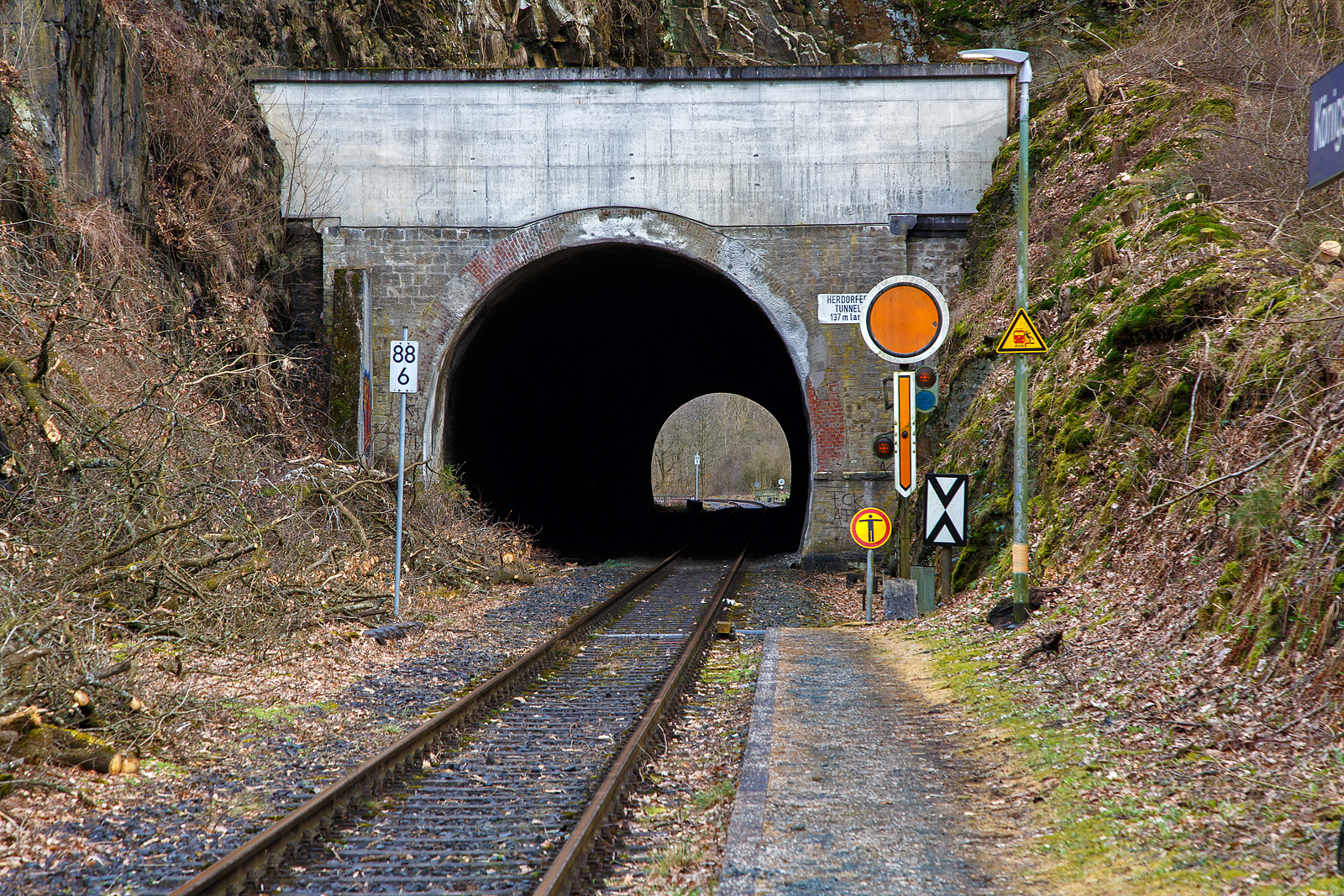 Der 137 m lange Herdorfer Tunnel hier am 18.03.2023 vom Hp Knigsstollen gesehen. 

Davor das Vorsignal (frs Einfahrtsignal Bf Herdorf) Vr 0 - Halt erwarten! Die gelbe Scheibe mit weiem Rand und gelbe Pfeil zeigt nach unten. Nachts leuchten auch die beiden gelben Lampen. Vor dem Signal die Ne 2 – Vorsignaltafel. Die Vorsignaltafel zeigt dem Lokfhrer, da das Signal, vor dem sie steht, Vorsignalfunktion hat, d.h., den Begriff eines nachfolgenden Hauptsignales vorankndigt. 
