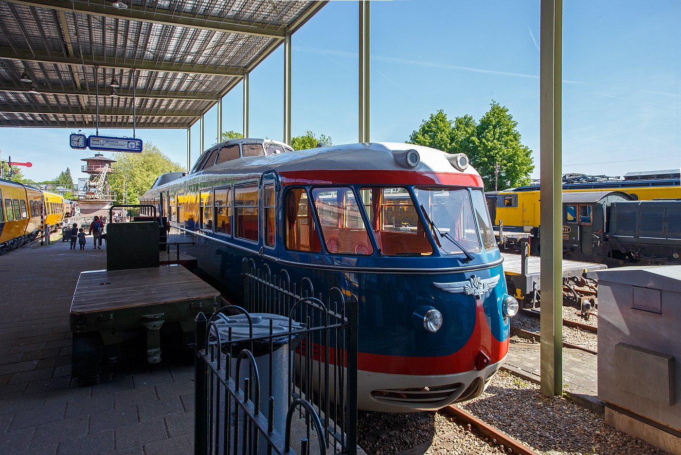 Der Dieseltriebwagen NS 20 „de Kameel“ / das Kamel bzw. VIP-Car, ein Dieseltriebwagen – Einzelstück und ehemaliger Inspektionstriebwagen, am 29 April 2025 im Het Spoorwegmuseum (bis 2005 NSM - Nederlands Spoorwegmuseum / Niederländischen Eisenbahnmuseum) in Utrecht-Maliebaan im Bahnhof Maliebaan. Ein sehr schönes Eisenbahnmuseum das zum Verweilen einlädt, leider fehlte uns dieses Mal die Zeit dafür. 

Der dieselelektrische Triebwagen wurde 1954 von Allan & Co´s Koninklijke Nederlandsche Fabrieken van Meubelen en Spoorwegmaterieel N.V. (Königlich Niederländische Möbel- und Eisenbahnausrüstungsfabrik) in Rotterdam gebaut und als Inspektionstriebwagen NS 20 an die NS - Nederlandse Spoorwegen (Niederländische Staatsbahnen) geliefert. Er diente für Inspektionsfahrten des NS-Vorstands.

Er technisch von den Triebwagen vom Typ DE 1 - Plan X „Blauwe Engel“ abgeleitet, nur der große Unterschied sind die Ausbuchtungen mit den Führerständen am Dach, die im bald den Spitznamen „Kamel“ einbrachten. 

1972 stand für den NS 20 eine Generalüberholung an. Zuvor durfte der Triebwagen in Toppop-Lackierung für die Musiksendung „Toppop“ durch die Niederlande touren. Im Rahmen der Überholung wurde der Triebwagen in Gelb umlackiert. Nach dieser Überholung stand der als „VIP-Wagen“ bekannte Triebwagen für Gruppenreisen zur Verfügung. Am 31. Januar 1991 fuhr der Wagen zum letzten Mal für die Öffentlichkeit, und am 8. März erfolgte die letzte Fahrt für die NS-Leitung. Anfang April fuhr die NS 20 mit eigenem Antrieb zur Lijnwerkplaats (Werkstatt) in Zwolle, um seine Generatoren als Ersatzteile zu spenden und sie der Reserve für die (inzwischen umgebauten und gelb lackierten) Blauwe-Engel-Züge hinzuzufügen. Diese Züge litten damals unter einem Mangel an Ersatzteilen.

Von diesem Zeitpunkt an war der NS 20 nicht mehr betriebsbereit und wurde zusammen mit einzelnen Dieselloks von der Lokomotive NS 2205 zum Eisenbahnmuseum transportiert und dort ausgestellt.

2005 wurde erneut ein Inspektionsfahrzeug benötigt, sodass eine Generalüberholung eingeleitet wurde, um das „Kamel“ wieder fahrbereit zu machen. Aus den ausgemusterten Plan X „Blaue Engel“ wurden Generatoren ausgebaut, zwei davon ausgewählt und nach Überholung wieder in den NS 20 eingebaut. Die Niederspannungsversorgung und die Küchenausstattung wurden in mehreren Bereichen für den modernen Einsatz modifiziert. Der in seiner originalen blauen Lackierung lackierte Triebwagen kehrte am 15. März 2008 mit eigener Kraft ins Museum zurück und wird dort betriebsbereit aufbewahrt.

Technik:
Auch die Frontpartie des Triebwagens anders als bei den DE 1 und 2. Das „Kamel“ ist 25.555 mm lang, somit fast 1,5 m kürzer als ein DE 1. Er hat eine Höhe von 4,145 Metern und eine Breite von 2,780 Metern. Das Gewicht des Fahrzeugs beträgt 58 Tonnen.

Der dieselelektrische Triebwagen verfügt über zwei Drehgestelle, die jeweils von zwei Elektromotoren angetrieben werden. Die elektrische Energie wird von zwei wassergekühlte AEC-6-Zylinder-Unterfur-Dieselmotoren Typ A220 (geliefert von Kemper & Van Twist), mit einer max. Leistung 132 kW (180 PS), Dauerleistung 107 kW (145 PS) mit gekoppeltem einem Generator von Smit vom Type G40/35 (147 kW) erzeugt, die jeweils zwei Heemaf-Fahrmotoren, Typ TM 51, antreiben. Die Dieselmotoren der DE 1 und 2 - Plan X „Blaue Engel“ hingegen haben durch einen Turbolader von Brown-Boveri eine höhere Dauerleistung von 165 kW (225 PS). Die maximale Drehzahl des Dieselmotors beträgt 1.250 U/min. Der Dieselmotor mit Generator, sowie Kompressor sind jeweils unter der Karosserie des Triebwagens montiert.

Der Triebwagen ist mit vier Heemaf-Fahrmotoren vom Typ TM 51 mit einer maximalen Leistung von 66 kW, 337 A bei 1.130 U/min und einer Dauerleistung von 49 kW, 248 A und 1.290 U/min ausgestattet. Die Energie liefert jeweils ein vom Dieselmotor angetriebener Generator für je 2 Motore. Kompressor und Hilfsgenerator werden über Keilriemen angetrieben. Diese befinden sich auf der anderen Seite des Hauptgenerators.

Der Radstand der Drehgestelle beträgt 3,0 Meter, der Mittenabstand zwischen den Drehgestellen 18,35 Meter. Der NS 20 ist für eine Höchstgeschwindigkeit von 125 km/h zugelassen. Der Raddurchmesser der Triebdrehgestelle, ist jedoch größer als bei den anderen Plan X-Garnituren, nämlich 1.250 mm. Die Drehgestelle sind jeweils mit zwei Heemaf-Fahrmotoren vom Typ TM 51 ausgestattet. Die Achsfolge ist somit Bo'Bo'. 

TECHNISCHE DATEN des DE 1 – 20 „de Kameel“:
Gebaute Anzahl: 1
Nummerierung: 20
Typ: dieselelektrischer Triebzug
Baujahr: 1954
Hersteller: Allan, Rotterdam
Spurweite: 1.435 mm (Normalspur)
Achsfolge: Bo'Bo' (4 Achsen in 2 Drehgestellen)
Länge über Kupplung: 25.550 mm
Drehzapfenabstand:
Achsanstand im Drehgestell: 3.000 mm
Breite: 2.780 mm
Höhe: 4.145 mm
Treibraddurchmesser: 1.250 mm (neu) 
Dieselmotor (2 Stück): wassergekühlte AEC-6-Zylinder-Unterfur-Dieselmotoren Typ A220
Motorhubraum: 11,31 Liter (Bohrung 130 mm x 142 mm Hub)
Motorleistung: 2 x 132 kW (180 PS)
Motordauerleistung: 2 x 107 kW (145 PS) bei 1.250 U/min
Generator (2 Stück): Smit G40/35 (147 kW)
E-Antriebsmotoren (4 Stück): Heemaf TM 51 max. 66 kW, 337 A bei 1.130 U/min
E-Motordauerleistung: 4 x 49 kW, 248 A und 1.290 U/min = 196 kW
Höchstgeschwindigkeit: 125 km/h
Eigengewicht: 58 t 

Quellen: Somda RailWiki.nl, Het Spoorwegmuseum, nmld.nl, Anschriften, Wikipedia (Niederlande)
Stand/Überarbeitung: September 2025
