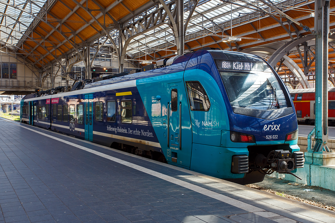 Der zweiteilige erixx Stadler FLIRT� Akku 526 022 / 826 022 (94 80 2526 022-5 D-SRSDE / 94 80 2826 022-2 D-SRSDE) im NAH.SH-Design (Schleswig-Holstein), als RB 84 L�beck Hbf nach Kiel Hbf, am 21 April 2026 im Hauptbahnhof L�beck. 

Der FLIRT wurde 2023 von Stadler Deutschland GmbH in Berlin- Pankow gebaut. Eigent�mer ist die Paribus Rail Portfolio III GmbH & Co. KG in Hamburg und vermietete diese an die erixx Holstein GmbH in L�beck, die die f�r den Nahverkehrsverbund Schleswig-Holstein (NAH.SH) einsetzt. Eingestellt diese FLIRT� Akku �ber die Stadler Rail Service Deutschland GmbH (D-SRSDE).

FLIRT Akku spart allein in Schleswig-Holstein j�hrlich 10 Millionen Liter Diesel und rund 26.000 Tonnen CO₂.

Mitte 2019 gewann Stadler eine Ausschreibung des Nahverkehrsverbunds Schleswig-Holstein �ber 55 FLIRT Akku f�r 600 Millionen Euro, einer der gr��ten Auftr�ge f�r Batteriez�ge in Deutschland. Seit Oktober 2023 ist diese weltweit erste batterieelektrische Zugflotte im regelm��igen Linienbetrieb unterwegs. Dank dieser Akkuz�ge hat das Bundesland den zweith�chsten Elektrifizierungsanteil unter den Fl�chenl�ndern Deutschlands, 68 Prozent aller Strecken werden nun elektrisch befahren. Der �kologische Vorteil: Rund 10,4 Millionen Zug-Kilometer werden „entdieselt“. 

Bald werden die FLIRT Akku-Triebz�ge in weiteren deutschen Bundesl�ndern wie auch in �sterreich, D�nemark, Litauen und den USA einen emissionsfreien Betrieb auf nicht elektrifizierten Strecken erm�glichen.

Der FLIRT Akku ist die batteriebetriebene Ausf�hrung der FLIRT-Typenreihe. Der Nahverkehrsverbund Schleswig-Holstein (NAH.SH) hat im Juli 2019 im Rahmen einer Innovationspartnerschaft 55 zweiteilige FLIRT Akku bestellt. Die batteriebetriebenen Triebz�ge verkehren seit Oktober 2023 auf den Strecken der Netze Nord und Ost.

Einsatzgebiete sind: Kiel-L�beck-L�neburg, Bad Oldesloe-Neum�nster, Neum�nster-Heide-B�sum, Kiel-Rendsburg-J�beck-Husum, Husum-Bad St.Peter Ording, Kiel-Eckenf�rde-Flensburg, Kiel-Sch�nberger Strand

Ausgelegt sind die FLIRT Akku Triebz�ge f�r nicht- oder nur teilelektrifizierte Strecken und so vielseitig einsetzbar. Das Laden der Batterien ist w�hrend der Fahrt unter Oberleitung sowie an elektrifizierten Endhaltepunkten m�glich. Zus�tzlich wird die Bremsenergie in den Batterien gespeichert. Damit kann das Fahrzeug effizient und energiesparend eingesetzt werden. Die Erbringung aller Instandhaltungs- und Serviceleistungen �ber 30 Jahre durch den Hersteller garantiert eine zuverl�ssige Verf�gbarkeit. Das mit Lithium-Ionen-Akkus ausgestattete Fahrzeug f�hrt in Schleswig-Holstein als zweiteilige Zugkomposition. Der voll klimatisierte FLIRT Akku f�r die NAH.SH verf�gt �ber mit Fahrgast-WLAN ausgestattete, helle Fahrgastr�ume sowie gro�z�gig gestaltete Mehrzweckbereiche und bietet 216 Fahrg�sten Platz, darunter 123 auf Sitzpl�tzen.

Technische Merkmale
Technik
Automatische Mittelpufferkupplungen
Leichtbauweise aus Aluminium
Luftgefederte Fahrwerke sorgen f�r die erforderliche Laufruhe
Oberleitungsbetrieb mit 15 kV sowie oberleitungsfreier Betrieb mit Lithium-Ionen-Traktionsbatterie
Schnellladung der Lithium-Ionen-Traktionsbatterie mittels 15 kV Oberleitung

Komfort
Heller, freundlicher Fahrgastraum
Klimatisierter Fahrgastraum und Fahrerraum
Gro�z�gig gestaltetes Multifunktionsabteil an allen Einstiegsbereichen
2 Einstiegst�ren pro Seite
Spalt�berbr�ckungen an allen T�ren
Fahrradstellpl�tze/Rollstuhlbereiche
Modernes Fahrgastinformationssystem
Universal WC nach TSI PRM
WLAN 

Personal
Ergonomisch gestalteter Fahrerarbeitsplatz

Spezifikationen
Betriebliche Reichweite im Akkubetrieb bis zu 150 km
H�chstgeschwindigkeit max. 160 km/h
Ladezeit unter Oberleitung: 15 Minuten
Sitzpl�tze: 123

Ersatzteillieferung und technische Instandhaltung �ber 30 Jahre durch Hersteller

FLIRT Akku: F�hrt da weiter, wo die Oberleitung endet
Moderne Z�ge m�ssen auf Strecken ohne Oberleitung nicht mit Diesel fahren. Leistungsstarke Batterien k�nnen Z�ge �ber Teilstrecken frei von CO₂-Aussto� und zuverl�ssig mit Strom versorgen. Die batterie-elektrischen Triebz�ge (BEMU) von Stadler sind ausgelegt f�r nicht- oder nur teilelektrifizierte Strecken und damit vielseitig einsetzbar.Auf Streckenabschnitten mit Oberleitung fahren die Z�ge klassisch elektrisch mit gehobenem Stromabnehmer und laden die Batterien. Gleichzeitig wird Bremsenergie zur Aufladung genutzt. Wo die Oberleitung endet, setzen sie ihre Fahrt im Batteriemodus fort. 

Mit dem FLIRT Akku, der batterieelektrischen Antriebsvariante des Bestsellers FLIRT, hat Stadler bereits 2017 den weltweit ersten f�r den Fahrgasteinsatz zugelassenen Batteriezug gebaut.

Das Fahrzeugkonzept beruht in gro�en Teilen auf den vielfach erprobten elektrischen Triebz�gen FLIRT f�r den Betrieb unter Oberleitung. Der FLIRT Akku ist einst�ckig in Leichtbauweise aus Aluminium gefertigt und in zwei- bis vierteiliger Zugkomposition realisierbar. In Serie l�sst sich der FLIRT Akku ebenso wie der FLIRT flexibel und individuell auf die jeweiligen Bed�rfnisse von Kunden und Streckennetz anpassen. Eine Mehrfachtraktion mit rein elektrischen ein- und doppelst�ckigen Fahrzeugen ist ebenfalls m�glich.

Die einzelnen Triebz�ge bestehen aus den zwei betrieblich nicht trennbaren Wagen z.B.:
Wagen A - 94 80 2826 022-2 D-SRSDE, 2. Klasse, Mehrzweckbereich
Wagen B - 94 80 2526 022-5 D-SRSDE, 1./2. Klasse, Rollstuhlpl�tze, barrierefreies WC;

TECHNISCHE DATEN:
Anzahl f�r NAH.SH: 55
Inbetriebsetzung Oktober 2023
Spurweite: 1.435 mm (Normalspur)
Achsanordnung: Bo´2´2´
L�nge �ber Kupplung: 45.700 mm
Fahrzeugbreite: 2.820 mm
Fahrzeugh�he: 4.275 mm
Achsabstand im Drehgestell: 2.700 mm
Trieb- und Laufraddurchmesser: 870 mm (neu)
Leergewicht: 96 t
H�chstgeschwindigkeit: 160 km/h
Dauerleistung am Rad: 1.000 kW (2 x 500 kW)
Traktionsantrieb von TSA: Motor TMF 59-39-4 und Getriebe GMH 2-43-595A
Speisespannung: 15 kV / 16,7 Hz
Stromversorgung: Stromabnehmer auf dem A-Wagen f�r 15 kV, 16,7 Hz AC aus der Oberleitung, 1 Batteriesystem im A-Wagen und 3 Batteriesysteme im B-Wagen
Batteriereichweite: 80 km laut Hersteller (bei Versuchsfahrten mehr als doppelt so weit)
Sitzpl�tze: 105 + 18 Klappsitze, davon 8 in der 1. Klasse
Stehpl�tze (4 Pers./m�): 123 (93 bei besetzten Klappsitzen)
Rollstuhlpl�tze: 2 im B-Wagen
Fahrradstellpl�tze: Im A-Wagen
Fu�bodenh�he: Niederflur 780 mm, Hochflur 1.145 mm
Einstiegsbreite: 1.300 mm
Einstiegsh�he: 780 mm

Quellen: Stadler Rail
