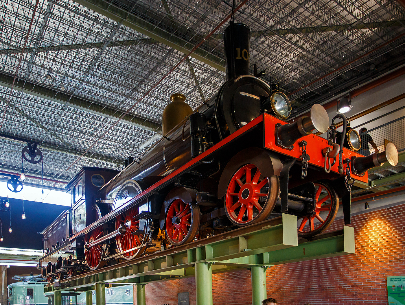 Die 1889 gebaute 2'B Schnellzug-Dampflokomotive NRS 107 „Rhijnboog“ (Rheinbogen), später HSM 353, NS 1604 (NS-Baureihe 1600), am 29 April 2025 hoch aufgeständert im Het Spoorwegmuseum (bis 2005 NSM - Nederlands Spoorwegmuseum / Niederländischen Eisenbahnmuseum) in Utrecht im ehemaligen Bahnhof Maliebaanstation.   

Die Dampflok wurde 1889 von Sharp, Stewart & Co. in Glasgow (GB) unter der Fabriknummer 3563 gebaut und an die NRS - Nederlandsche Rhijnspoorweg-Maatschappij (Niederländische Rheinbahn-Gesellschaft). Im Jahr 1890 wurde die NRS vom niederländischen Staat übernommen, der ihren Betrieb an die Staatsbahnen (SS) übertrug. Das NRS-Rollmaterial wurde zwischen den Staatsbahnen (SS) und der Hollandse IJzeren Spoorweg-Maatschappij (HSM) aufgeteilt, so wurde die NRS 107 zur HSM 353. Ab 1921, nach der Zusammenlegung des Rollmaterials der SS und der NRS, wurde die Lok zur NS 1604.

Die Baureihe NS 1600 war eine Reihe von 59 Schnellzugdampflokomotiven der NS - Nederlandse Spoorwegen (Niederländischen Eisenbahnen) und ihrer Vorgängergesellschaften, der NRS - Nederlandsche Rhijnspoorweg-Maatschappij (Niederländische Rheinbahn-Gesellschaft), der SS - Maatschappij tot Exploitatie van Staatsspoorwegen (Gesellschaft zur Nutzung der Staatseisenbahnen) und der HSM - Hollandsche IJzeren Spoorweg-Maatschappij (Niederländischen Eisenbahn-Gesellschaft). Die Loks wurden von Sharp, Stewart & Co. in Glasgow (GB) gebaut.

Die Loks vom Typ „Rhijnboog“ stellt einen großen Sprung nach vorn im Lokbau dar. Es war die erste Lokomotive, die vorne ein führendes Lauflachsen-Drehgestell hatte, das die Fahrleistung verbesserte. Dies ermöglichte auch die Länge und damit die Kapazität des Kessels zu erhöhen. Sie gehörten damals zu den größten Lokomotiven Europas.

Der Spitzname  Rhijnboog  kombiniert den Namen der niederländischen Eisenbahngesellschaft, die die Lokomotive (Nederlandsche Rhijnspoorweg-Maatschappij) mit dem Wort  Bogie  bestellt hat. Nach der Auflösung der NRS wurde die Lokserie schließlich von der HSM übernommen. Sie waren mit diesen Loks so zufrieden, dass sie 50 weitere nahezu identische Lokomotiven bestellte. Die Lokomotive NRS 107 hat einen besonderen Platz im Museum, sie überragt die Besucher von seinem erhabenen  Thron . 

NRS 101 bis 109:
Im Jahr 1889 bestellte die Nederlandsche Rhijnspoorweg-Maatschappij (NRS) neun Lokomotiven mit den Nummern 101–109 bei der Fabrik von Sharp Stewart and Company in Glasgow. Die Achsfolge der Lokomotiven war 2'B. Zum Zeitpunkt der Bestellung waren dies die ersten niederländischen Lokomotiven mit einem Laufdrehgestell vor den Antriebsrädern (Triebachsen). Sie waren die letzten von der NRS bestellten Lokomotiven. Ein Jahr später, 1890, wurde die NRS aufgelöst und die Ausrüstung zwischen der Niederländischen Eisenbahn-Gesellschaft (HSM) und der Staatlichen Eisenbahn-Ausbeutungsgesellschaft (SS) aufgeteilt. Die Lokomotiven 101, 102, 103, 108 und 109 gingen an die SS und erhielten die SS-Nummern 1101–1103 und 1108–1109. Die 104-107 gingen an das HSM und bekamen die HSM-Nummern 350-353. Da die Lokomotiven für die kleinen Drehscheiben der SS zu lang waren, übergab die SS die fünf Lokomotiven ein Jahr später, 1891, an die HSM, wo sie die Nummern 354–358 erhielten. Aufgrund des Drehgestells für die Antriebsräder, auch Bogie genannt, und der Herkunft des Rhijnspoor erhielten diese Maschinen bei der HSM den Spitznamen „Rhijnboog“.

HSM 359 bis 408:
Die HSM war mit den von ex NRS Lokomotiven so zufrieden, dass sie 40 weitere bei Sharp Stewart and Company bestellte. Diese wurden zwischen 1891 und 1892 unter den Nummern 359–398 in Dienst gestellt. Im Jahr 1900 wurde eine weitere Bestellung über 5 Einheiten mit den Nummern 399-403 bei Sharp Stewart and Company aufgegeben. Schließlich wurde im Jahr 1903 die Klasse 404–408 in Betrieb genommen, die von der North British Locomotive Company (Atlas Works) gebaut wurden, da Sharp Stewart & Co. Company inzwischen zu dieser Gesellschaft fusioniert war.

Bedingt durch den Ersten Weltkrieg wurde 1917 eine Zweckgemeinschaft der unabhängigen Eisenbahngesellschaften HSM (Hollandsche IJzeren Spoorweg-Maatschappij) und SS (Maatschappij tot Exploitatie van Staatsspoorwegen) gegründet. Die beiden Gesellschaften errichteten einen gemeinsamen Bahnbetrieb, blieben aber rechtlich unabhängig. Dies änderte sich 1938, als HIJSM und SS vollständig fusionierten und in der AG NS (Nederlandse Spoorwegen, früher Nederlandsche Spoorwegen geschrieben) aufgingen. Das Rollmaterial wurde bereits 1921 zusammengelegt, so wurden die Lok zur NS 1604 nummeriert.

Die NS 1614, 1633, 1636, 1637 und 1646 wurden 1921 mit einem Überhitzer ausgestattet. Es war beabsichtigt, die gesamte Serie damit auszurüsten, aber aufgrund enttäuschender Ergebnisse blieb es bei diesen fünf Lokomotiven. Sie wurden in den Jahren 1936–1949 außer Dienst gestellt. Die Lokomotive NS 1622 wurde als letzte dieser Baureihe im Jahr 1949 ausgemustert. Die NS 1604, ex HSM 353, ex NRS 107 „Rhijnboog“ blieb bis 1940 im Einsatz blieb. Die 1604 wurde zur Erhaltung für das Niederländische Eisenbahnmuseum in Utrecht ausgewählt und im Lokschuppen in Maastricht eingelagert, wo die Lokomotive am 8. September 1944 durch Kriegseinwirkung schwer beschädigt wurde. Nach dem Zweiten Weltkrieg wurde die Lokomotive von der NS-Hauptwerkstatt in Tilburg vollständig restauriert und in die braune Version der NRS mit der ursprünglichen Nummer 107 zurückversetzt. Seit März 1955 ist die NRS 107 im Eisenbahnmuseum ausgestellt. Nach der Sanierung des Eisenbahnmuseums in den Jahren 2003 bis 2005 wurde die Lokomotive aufgeständert.	

TECHNISCHE DATEN:
NRS-Serie: Rhijnboog, ab 1921 NS 1600
Hersteller: Sharp, Stewart & Co. in Glasgow (GB), später Atlas Works
Gebaute Anzahl: 59
Spurweite: 1.435 mm (Normalspur)
Achsfolge: 	2'B (engl. 4-4-0), gekuppelt mit dreiachsigem Tender
Länge über Puffer: 16.585 mm 
Treibraddurchmesser: 2.016 mm (neu) 
Laufraddurchmesser (Lok und Tender): 1.105 mm (neu) 
Breite: 2.222 mm
Höhe: 4.267 mm
Höchstgeschwindigkeit: 90 km/h
Rostfläche: 2,15 m²
Gesamtheizfläche: 111,6  m²
Kesselüberdruck: 10,3 kg/cm²
Anzahl der Zylinder: 2 (Ø 457 mm x 660 mm Hub)
Steuerung: Stephenson
Eigengewicht: 81,3 t (Lok 48,8 t und Tender 32,5 t) 
Anhängelast: 4.950 kg
Wasservorrat: 10,2 m³ 
Kohlevorrat: 2,8 t
