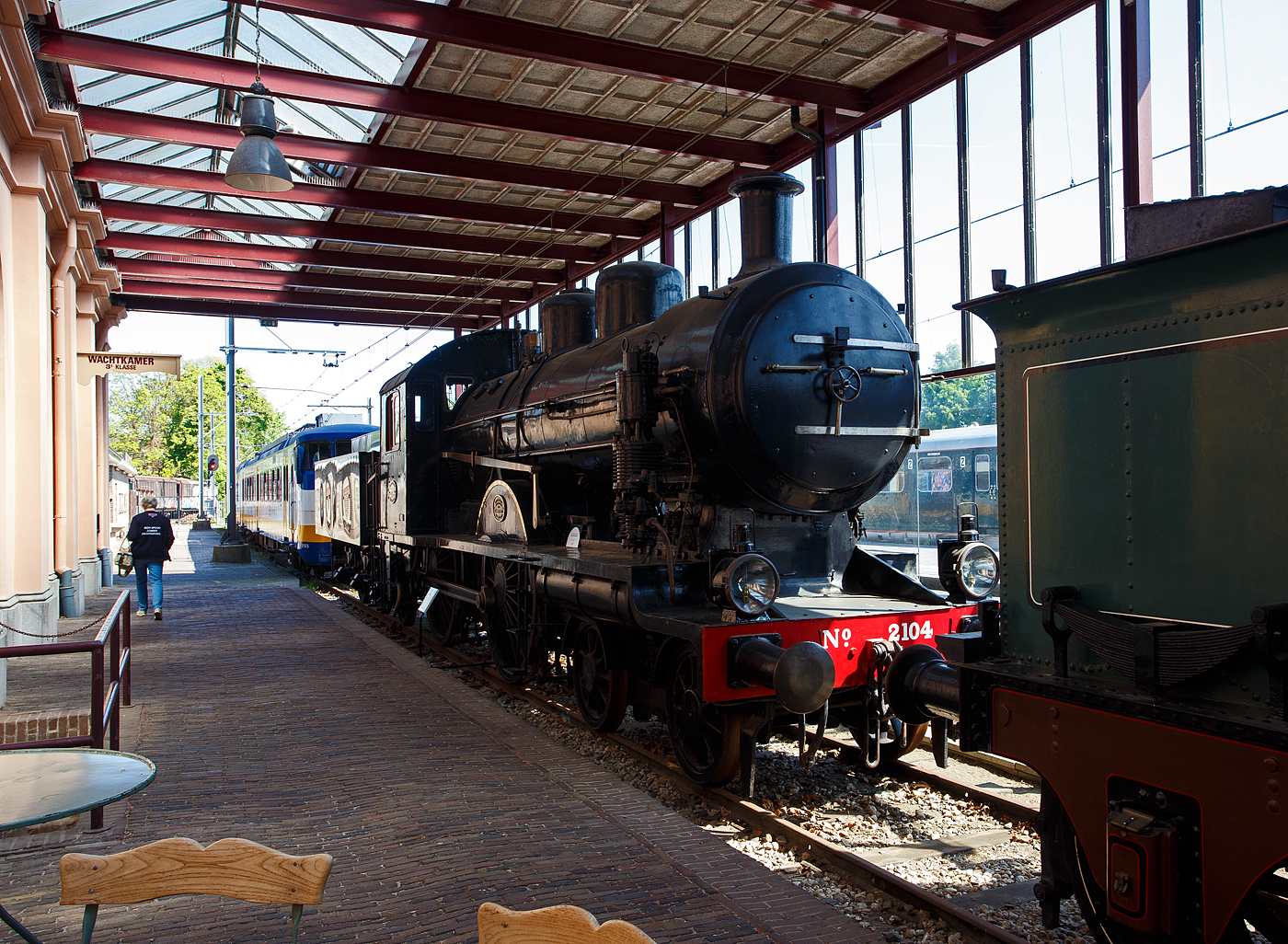 Die 1914 gebaute 2'B Schnellzug-Dampflokomotive NS N° 2104 „Blikken Tinus“ (blecherner Martin) der NS-Baureihe 2100, ex HSM 504, am 29 April 2025 im Het Spoorwegmuseum (bis 2005 NSM - Nederlands Spoorwegmuseum / Niederländischen Eisenbahnmuseum) in Utrecht im Bahnhof Maliebaanstation.   

Die Dampflok wurde 1914 von der BMAG - Berliner Maschinenbau-Actien-Gesellschaft vormals L. Schwartzkopff in Berlin unter der Fabriknummer 5304 gebaut und als HSM 504 an die Hollandse IJzeren Spoorweg-Maatschappij (Holländische Eisenbahn-Gesellschaft) geliefert. 

Die Lokomotiven der Baureihe NS 2100 waren Schnellzugdampflokomotiven der Nederlandse Spoorwegen (NS), die von der Hollandsche IJzeren Spoorweg-Maatschappij (HSM) übernommen wurden.

Die Lokomotiven hatten die Achsfolge 2’B. Die Serie wurde vom HSM-Ingenieur Willem Hupkes für die Beförderung der immer schwereren Personen- und Schnellzüge konzipiert. Die ersten 5 Loks (HSM 501 – 505) wurden 1914 von der BMAG in Deutschland gebaut, 1916 bis 1919 folgten 15 Stück (516 – 530) von Werkspoor in Amsterdam (NL), 1920 folgten 10 Stück (506-515) von der BMAG aus Deutschland, sowie 5 Stück (531 – 535) von Werkspoor aus Amsterdam.

Da das Konkurrenzunternehmen der HSM, die SS - Maatschappij tot Exploitatie van Staatsspoorwegen (Gesellschaft zur Nutzung der Staatseisenbahnen) zu stärkeren 2’C-Maschinen der Serie SS 700 übergegangen war, konnte die HSM nicht zurückbleiben. Obwohl ein HSM-Entwurf für eine 2’C-Maschine bekannt ist, wurde letztlich eine 2’B-Lokomotive ausgewählt. Dies lag jedoch nicht an der begrenzten Länge der Drehscheiben, wie oft fälschlicherweise angenommen wird. Die Maschinen hatten jedoch die maximal zulässige Achslast von 17 Tonnen und die größten Antriebsräder (Ø 2.100 mm) in den Niederlanden, weshalb sie ideal für Schnellzüge waren. Wegen des rasselnden Geräuschs der Kuppelstangen wurden sie vom Personal „Blikken Tinus“ – deutsch blecherner Martin – genannt. Die Lokomotiven wurden von der Berliner Maschinenbau-AG vormals L. Schwartzkopff (BMAG) in Berlin und Werkspoor in Amsterdam gebaut.

Bedingt durch den Ersten Weltkrieg wurde 1917 eine Zweckgemeinschaft der unabhängigen Eisenbahngesellschaften HSM (Hollandsche IJzeren Spoorweg-Maatschappij) und SS (Maatschappij tot Exploitatie van Staatsspoorwegen) gegründet. Die beiden Gesellschaften errichteten einen gemeinsamen Bahnbetrieb, blieben aber rechtlich unabhängig. Dies änderte sich 1938, als HIJSM und SS vollständig fusionierten und in der AG NS (Nederlandse Spoorwegen, früher Nederlandsche Spoorwegen geschrieben) aufgingen. Das Rollmaterial wurde bereits 1921 zusammengelegt, so wurden die Lok zur NS 2104 nummeriert. Die Lokomotive 2104, ehemals HSM 504, wurde als letzte dieser Baureihe 1954 ausgemustert und blieb im Nederlands Spoorwegmuseum erhalten.

TECHNISCHE DATEN:
Hersteller: BMAG in Berlin und Werkspoor in Amsterdam
Baujahre: 1914 bis 1920
Gebaute Anzahl: 35
Spurweite: 1.435 mm (Normalspur)
Achsfolge: 	2'B (engl. 4-4-0), gekuppelt mit dreiachsigem Tender
Länge über Puffer: 17.845 mm 
Treibraddurchmesser: 2.100 mm (neu) 
Laufraddurchmesser (Lok): 990 mm (neu) 
Laufraddurchmesser (Tender): 1.105 mm (neu)
Höhe: 4.370 mm
Höchstgeschwindigkeit: 110 km/h
Dienstgewicht: 105 t (61 t Lok und 44 t Tender)
Anfahrzugkraft: 7.660 kg
Rostfläche: 2,4 m²
Gesamtheizfläche: 154 m² (Strahlungs- 12 m², Rohr- 107 m² und Überhitzer- 35 m²
Kesselüberdruck: 12,4 kg/cm²
Anzahl der Zylinder: 2 (Ø 530 mm x 660 mm Hub)
Steuerung: Walschaerts
Wasservorrat: 19 m³ 
Kohlevorrat: 6,0 t
