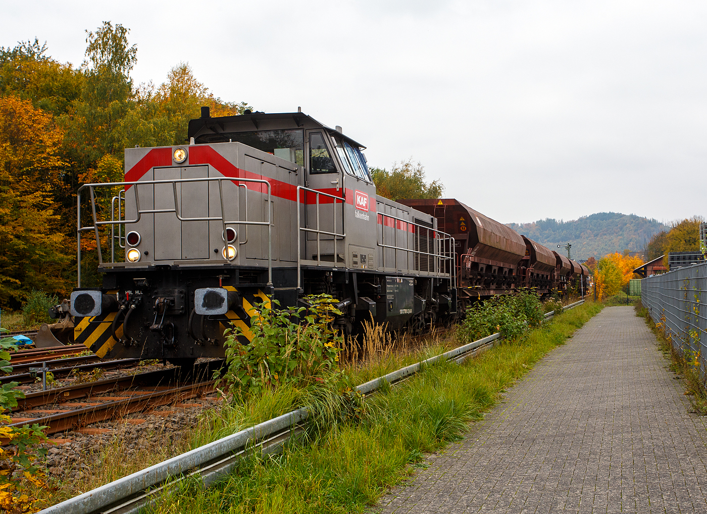 Die 277 809-0  Elmi“ (92 80 1277 809-0 D-KAF), eine Vossloh MaK G 1700 BB der KAF - Falkenhahn Bau AG (Kreuztal), steht am 12 Oktober 2025 mit einem Schotterzug im Bahnhof Herdorf. Mit dem Zug werden später die neuen Weichen 25 und 26 eingeschottert. 

Die Vossloh MaK G 1700 BB wurde 2001 von Vossloh in Kiel 2001 unter der Fabriknummer 1001113 gebaut und an die NE - Neusser Eisenbahn geliefert. Nach dem Zusammenschluss der HGK und der NE zur RheinCargo GmbH & Co. KG, fuhr sie noch als RHC 277 809-0. Anfang 2014 ging sie an die Vossloh Locomotives GmbH (92 80 1277 809-0 D-VL) und war 1 ½ Jahre als Mietlok unterwegs, bis sie im Juni 2015 an die Michael Bugdoll Bau- und Sicherungsarbeiten e. K in Dorsten ging, als diese konnte ich sie auch mal ablichten. Seit 01.01.2021 ist die Lok nun bei der KAF - Falkenhahn Bau AG (Kreuztal), hier hat sie aber ihren Namen  Elmi“ behalten. 

TECHNISCHE DATEN:
Baujahr: 2001 - 2003
gebaute Stückzahl: 6
Spurweite: 1.435 mm (Normalspur)
Achsfolge: B´B´
Länge über Puffer: 14.700 mm
Drehzapfenabstand: 7.400 mm
Achsabstand im Drehgestell: 2.400 mm
größte Breite: 3.080 mm
größte Höhe über Schienenoberkante: 4.220 mm
Raddurchmesser: 1.000 mm (neu)
kleinster befahrbarer Gleisbogen: 60 m
Dienstgewicht 87,3 t
Kraftstoffvorrat 3.500 l
Motor: MTU-12-Zylinder-Dieselmotor, vom Typ 12V 4000 R20
Leistung: 1.500 kW bei 1800 U/min
Getriebe: Voith L 5r4 zU2
Anfahrzugkraft: 249 kN
Höchstgeschwindigkeit: 100 km/h

Die Vossloh Lokomotive MaK G 1700 BB ist eine dieselhydraulische Lokomotive. Bei der Überarbeitung der Typenpalette nach der Übernahme der MaK durch Vossloh 1998 wurde im 4. Typenprogramm unter der Bezeichnung G 1700 BB der Bau einer vierachsigen Streckenlokomotive mit Mittelführerhaus vorgesehen. Tatsächlich handelte sich zunächst um eine Weiterentwicklung der G 1206. Die Maschine wurde in einigen Details an die Lokomotiven des 4. Typenprogramms angepasst. Dies betraf in erster Linie den Rahmen, der so geändert wurde, dass die neuen Standarddrehgestelle verwendet werden konnten. Dabei wurden die Rahmenenden so umgestaltet, dass die Lokomotiven die im 4. Typenprogramm üblichen Komfortaufstiege erhielten. Ebenfalls geändert wurde der Dieselmotor. Zum Einsatz kam der schon bei den G 1206 für die Dortmunder Eisenbahn verwendete MTU 12V 4000 R20 mit einer unveränderten Leistung von 1.500 kW bei 1.800 U/min. Die übrigen Komponenten der G 1700 BB blieben gegenüber der G 1206 weitgehend unverändert.

Der Dieselmotor ist ein wassergekühlter MTU 90°-V12-Zylinder-Viertakt-Dieselmotor mit Common-Rail-Einspritzung, OHV-Ventilsteuerung, Registeraufladung mit Ladeluftkühlung. Der Hubraum beträgt 57.199 cm³ (57,2 Liter), die Zylinder haben eine Bohrung von Ø 170 mm × 210 mm Hub. Die Motorabmessungen (LxBxH) sind 2.670 x 1.696 x 2.001 mm, das Gewicht (trocken) beträgt 7.700 kg.

Die ersten beiden Lokomotiven dieses Typs (die KSW Lok 42 und diese hier) wurden im Jahr 2001 ausgeliefert. Ende 2002 bis Anfang 2003 folgten weitere vier Lokomotiven aus Vorratsbauten. Nach diesen sechs Lokomotiven wurde der Bau der G 1700 BB zugunsten der technisch sehr ähnlichen und bereits weiter verbreiteten G 1206 wieder eingestellt.

So ist der Nachfolger die G 1700-2 BB keine Weiteentwicklung der G 1700 BB, sondern um eine komplette Neuentwicklung. 
