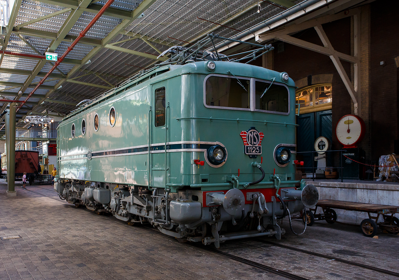 Die Elektrolokomotive NS 1125 der NS-Baureihe 1100 am 29 April 2025 im Het Spoorwegmuseum (bis 2005 NSM - Nederlands Spoorwegmuseum / Niederländischen Eisenbahnmuseum) in Utrecht im ehemaligen Bahnhof Maliebaanstation. Ein Eisenbahnmuseum das zum Verweilen einlädt, leider fehlte uns dieses Mal die Zeit dafür.  

Die Lok wurde 1951 von Alstom in Belfort (Frankreich) gebaut und an die NS - Nederlandse Spoorwegen (Niederländischen Staatsbahnen, heute Niederländische Eisenbahnen AG), sie war Teil der Serie NS 1100 von 60 Stück vierachsiger Elektrolokomotiven.

Die 60 Lokomotiven der Baureihe 1100, die zwischen 1950 und 1956 von der NS in Dienst gestellt wurden, waren für D-Züge, Schnellzüge und Güterzüge, sowie zunächst auch für TEE-Züge vorgesehen. Die Loks basierte auf der vierachsigen SNCF-Lokomotive der Serie BB 8100. Die französischen Lok BB 8100 waren für den Güterverkehr vorgesehen und hatten eine Höchstgeschwindigkeit von 105 km/h. In der Praxis fuhren sie nicht schneller als etwa 80 km/h. Allerdings mussten die niederländischen 1100er über längere Strecken mit 130 km/h fahren, wofür die Konstruktion der Drehgestelle drastisch geändert wurde. Die Höchstgeschwindigkeit betrug so 130 km/h. Alsthom lieferte die 1101 am 3. Juli 1950 als erstes Unternehmen an die NS-Hauptwerkstatt in Tilburg. 

Die niederländischen 1100er ähneln stark den französischen BB 8100er. Sie unterscheiden sich neben der Optik vor allem durch stärkere Motoren, eine Federung mit einer Kombination aus Federn und Gummiblöcken, Rundpuffern und einem rechts angebrachten Fahrerpult. Ihre Höchstgeschwindigkeit liegt mit 12´35 km/h über der der BB 8100.

Ursprünglich war die Hauptfarbe der Lokomotiven (1101 – 1150) türkis mit einer roten Pufferbohle und grauen Drehgestellen. Da Türkis zu leicht Schmutz zeigte, wurde die Hauptfarbe ab 1954 allmählich durch dunkelblau (preußisches Blau) ersetzt. Die 1151 – 1160 wurden direkt in Blau mit schwarzen Drehgestellen ausgeliefert. Die Pufferbohle blieb rot.

Ab 1971 wurden die Lokomotiven in der damals üblichen gelbgrauen Lackierung lackiert. Bei der 1125 geschah dies erst im Jahr 1980. Im selben Jahr erhielt die Lokomotive eine Bugnase, die den Lokführer bei Kollisionen besser schützen sollte. Die Drehgestelle wurden zudem verlängert. Die 1125 wurde am 27. September 1987 außer Dienst gestellt und ins Eisenbahnmuseum gebracht. Im Jahr 1989 wurde, als Teil der Feierlichkeiten zum 150. Jahrestag der niederländischen Eisenbahn, die 1125 restauriert und ihre Nasenkappe entfernt und in ihren ursprünglichen Zustand zurückversetzt.

Der 1100ter war als echter Allrounder bekannt, auf den man sich verlassen konnte, um sowohl Personenzüge als auch Güterzüge zu ziehen, ob leicht oder schwer. Allerdings waren die Fahreigenschaften eher mittelmäßig, so wurde viel Aufwand in die Verbesserung gesteckt, so richtig gut wurden sie aber nie.

Crashnase (Botsneus):
Die NS 1129 kollidierte 1978 bei Westervoort mit einem Dieseltriebzug. Diese wurde wieder repariert. Bei der Restaurierung der Lokomotive ging ein lang gehegter Wunsch der Lokführer in Erfüllung. Hinter der geraden Front fühlten sich die Lokführer nicht sehr sicher. Die flache Front wurde um eine Knautschzone in Form einer Frontnase, der sogenannten Crashnase, erweitert (es wurde keine Beplankung entfernt), um dem Fahrer im Falle eines Aufpralls mehr Schutz zu bieten. Bis 1981 wurden alle noch vorhandenen 58 Lokomotiven der Serie 1100 auf diese Weise umgebaut.

TECHNISCHE DATEN:
Hersteller: Alsthom, Belfort (Frankreich)
Baujahre: 1950 bis 1956
Ausmusterung: bis 1999
Gebaute Anzahl: 60
Nummerierung: NS 1101 bis 1160
Spurweite: 1.435 mm (Normalspur)
Achsfolge: Bo’Bo’ 
Länge über Puffer: 12.984 mm ursprünglich (flache Front) / 14.184 mm mit Crashnase
Drehzapfenabstand: 6.000 mm
Achsabstand im Drehgestell: 2.950 mm
Treibraddurchmesser: 1.250 mm (neu)
Gewicht: 80 t ursprünglich / 83 t mit Crashnase
Antriebsmotoren: 4 Stück vom Typ Alsthom TA 628 A
Getriebeübersetzung: 20 : 74
Höchstgeschwindigkeit: 130 km/h 
Stundenleistung: 2.290 kW (3.115 PS)
Dauerleistung: 2.050 kW (2.287 PS)
Anfahrzugkraft: 155 kN
Dauerzugkraft: 102 kN
Stromsystem: 1,5 kV DC (=)
Kleister befahrbarer Gleisbogen: R 80 m
