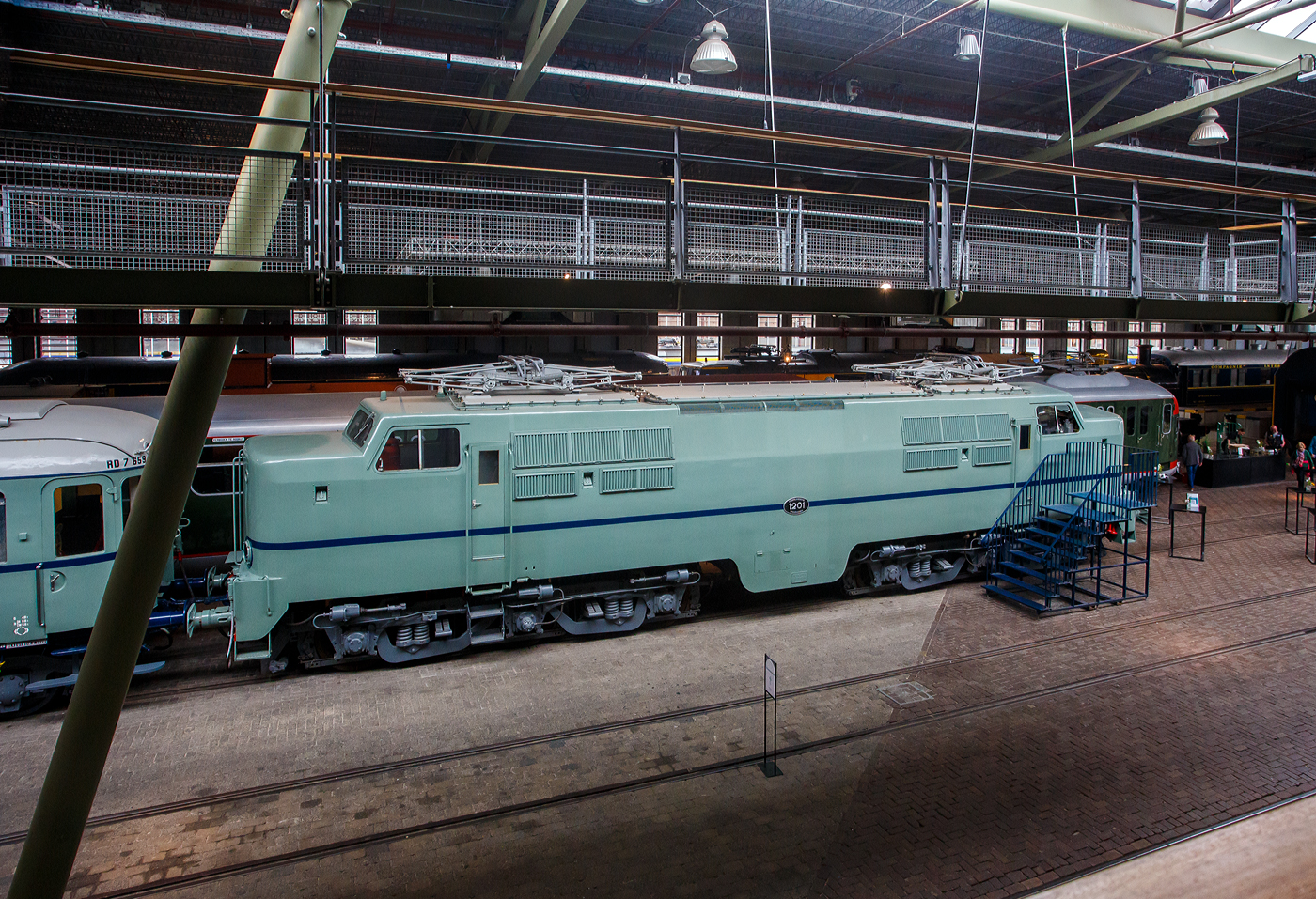 Die Elektrolokomotive NS 1201 der NS-Baureihe 1200 am 24 Juni 2025 im Het Spoorwegmuseum (bis 2005 NSM - Nederlands Spoorwegmuseum / Niederländischen Eisenbahnmuseum) in Utrecht im ehemaligen Bahnhof Maliebaan. Ein Eisenbahnmuseum das zum Verweilen einlädt.

Die Lok wurde 1951 von Werkspoor in Utrecht unter der Fabriknummer 928 gebaut (Baldwin-Lizenz) und an die NS - Nederlandse Spoorwegen (Niederländischen Staatsbahnen, heute Niederländische Eisenbahnen AG) geliefert. Wobei die Entwicklung und Konstruktion, sowie auch einige Teile, von Baldwin Locomotive Works in Philadelphia (USA) stammt. Auch die Entwicklung der Elektrotechnik stammt aus den Vereinigten Staaten und zwar von Westinghouse Electric Corporation (Monroeville), diese wurde von Heemaf in Hengelo (Westinghouse-Lizenz) geliefert.

Die Baureihe 1200 der Niederländischen Eisenbahnen (NS) ist eine sechsachsige Elektrolokomotivbaureihe aus den Jahren 1951 bis 1953. Die Lokomotiven waren bis 1998 bei der NS im Einsatz.

Die Nase war der erste Teil dieser unglaublich starken und robusten Lokomotive, der ins Auge fiel. Sie sollte dem Lokführer bei Kollisionen einen besseren Schutz bieten. Diese Lokomotiven wurden in den Niederlanden nach einem amerikanischen Design und mit einigen amerikanischen Teilen gebaut, die teilweise durch amerikanische Dollar über den Marshall-Plan finanziert wurden. Die NS nutzte diese Lokomotiven, um sowohl Personenzüge als auch Güterzüge zu ziehen. Ihre wichtigste Aufgabe war es Schnellzüge zu ziehen. Als sich die NS 1995 trennte, wurden alle 1200er Jahre an NS Cargo übertragen und begannen im Güterverkehr zu arbeiten. 

Nach Ende des Zweiten Weltkrieges gab es großen Fahrzeugmangel bei der NS. Da man keine Erfahrungen mit dem Bau elektrischer Lokomotiven hatte, kaufte man Lokomotiven bei verschiedenen Lieferanten. Die bei Baldwin konstruierten Lokomotiven wurden bei Werkspoor in Utrecht gefertigt. Die bei Westinghouse entwickelte Elektrotechnik wurde von Heemaf in Hengelo geliefert. Von den ursprünglich 75 georderten Lokomotiven wurden nur 25 ausgeliefert. Der amerikanische Einfluss ist an den hochliegenden Führerständen und den großen, dem Schutz des Lokführers dienenden Vorbauten erkennbar. Am 30. November 1950 wurden 25 Loks bestellt, am 27. September 1951 konnte die erste Probefahrt stattfinden. Am 9. Februar 1952 wurde die 1201 offiziell in Dienst gestellt, der Preis pro Lok betrug 765.000 NFL (ca. 347.000 Euro).

Die Loks hatten sechs Motoren des Typs TM94. Die ersten 14 Lokomotiven waren türkis lackiert, danach setzte sich wegen des sichtbaren Abriebs der Stromabnehmer eine rotbraune Farbgebung durch. Ab 1954 erhielten alle Loks eine blaue Farbgebung, 1971 erhielt die erste Lok die neuen NS-Hausfarben grau/gelb, 1983 war auch die letzte Lok entsprechend umlackiert. Am 30. März 1998 endete der planmäßige Einsatz bei der NS.

Fünf weitere Lokomotiven (1214, 1215, 1218, 1221, 1225) wurden 1999 an die ACTS Nederland BV verkauft. Diese vermietete 2010 drei Lokomotiven (1214, 1215, 1225) an die Eisenbahngesellschaft Euro-Express Treincharter (EETC). Von der wurden sie bis April 2015 unter anderem vor den Nachtzügen Niederlande–Österreich zwischen Amsterdam und Venlo eingesetzt. Fünf Maschinen (1201, 1202, 1211, 1218, 1221) sind als Museumslokomotiven erhalten. NS 1211 ist Botschafterlok im Bahnpark Augsburg. Leihgeber ist das Eisenbahnmuseum Utrecht.

TECHNISCHE DATEN:
Hersteller mech.: Werkspoor (in Lizenz von Baldwin)
Hersteller Elektrik: Heemaf (in Lizenz von Westinghouse)
Baujahre: 1950 bis 1953
Ausmusterung bei NS: bis 1998 
Gebaute Anzahl: 25
Nummerierung: NS 1201 bis 1225
Spurweite: 1.435 mm (Normalspur)
Achsfolge: Co’Co’ 
Länge über Puffer: 18.086 mm
Höhe: 4.064 mm
Treibraddurchmesser: 1.100 mm (neu)
Dienstgewicht: 108 t
Antriebsmotoren: 6 Stück Heemaf vom Typ TM 94
Getriebeübersetzung: 20 : 71
Höchstgeschwindigkeit: 130 km/h (Planmäßig) / 150 km/h (technisch möglich)
Stundenleistung: 6 x 392 kW = 2.350 kW
Dauerleistung: 6 x 367 kW = 2.200 kW
Anfahrzugkraft: 195 kN
Dauerzugkraft: 164 kN bei 80 km/h / 64,7 kN bei 123 km/h
Stromsystem: 1,5 kV DC (=)
Kleister befahrbarer Gleisbogen: R 100 m