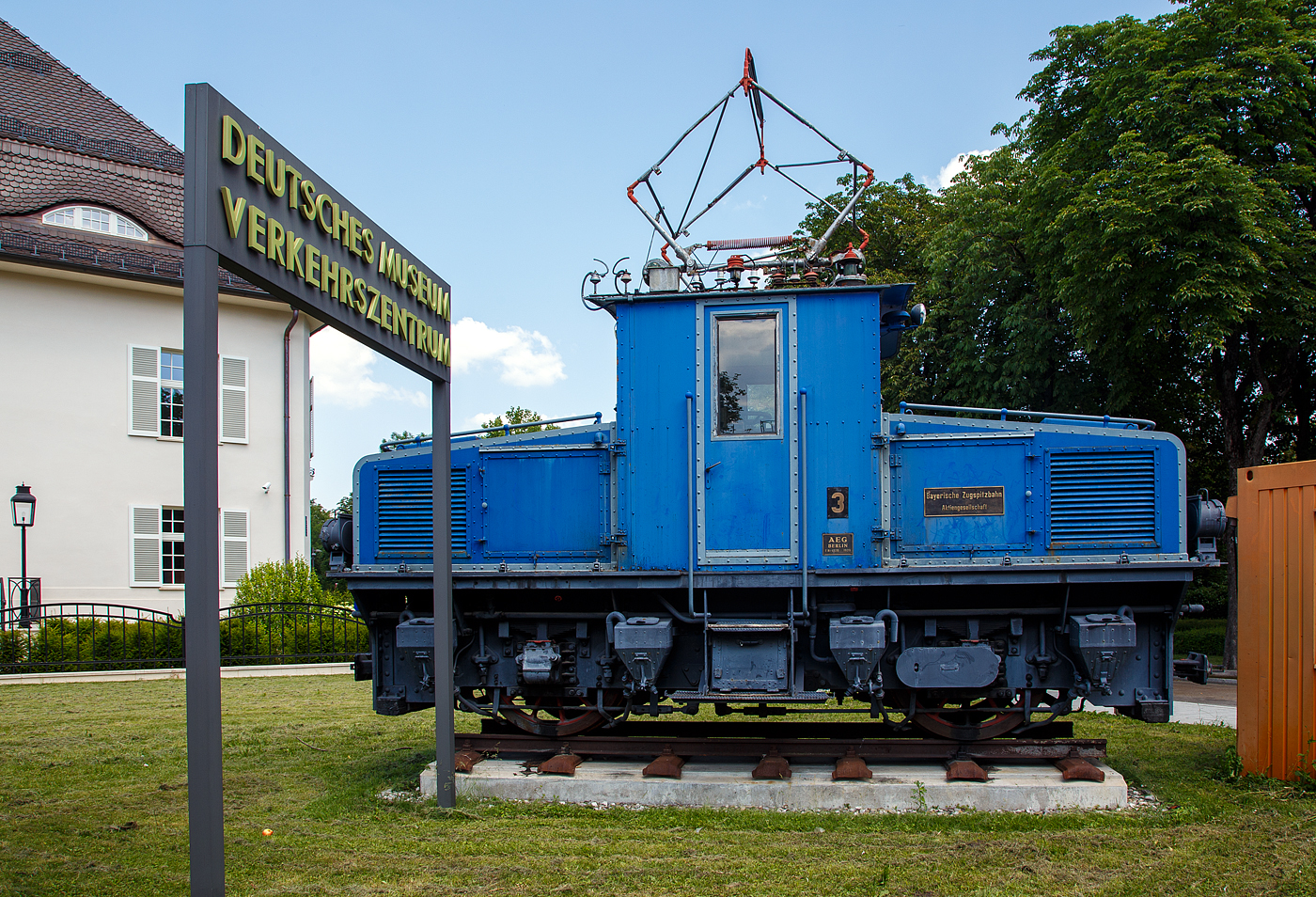 Die Tallokomotive 3 (Adhäsionslok) der Bayerische Zugspitzbahn AG als Denkmal beim Verkehrszentrum des Deutschen Museums in München am 16.06.2018. 

Die Lok wurde 1929 von AEG in Berlin unter der Fabriknummer 4270 gebaut. Seit April 2008 steht sie zusammen mit der Berglokomotiven 11 (Zahnradlokomotive) als Denkmal vor dem Verkehrszentrum des Deutschen Museums in München, sie sind eine Leihgabe der BZB ans Museum.

Die Tallokomotiven 1 ist auch noch im Jahr 2020 noch im Betriebsbestand und wird für Rangieraufgaben im Betriebswerk Grainau oder vor Bauzügen auf der Talstrecke eingesetzt. 

Die Tallokomotiven der Bayerischen Zugspitzbahn sind meterspurige Elektrolokomotiven für den reinen Adhäsionsbetrieb. Sie wurden zusammen mit den Berglokomotiven 11 bis 18 als Erstausstattung der Bayerischen Zugspitzbahn 1929 beschafft. Die zweiachsigen Tallokomotiven für die Talstrecke zwischen Garmisch-Partenkirchen und Grainau mit den AEG Fabriknummern 4268 bis 4271 hatten die Aufgabe, maximal sechs Personenwagen mit bis zu 40 km/h von Garmisch-Partenkirchen nach Grainau zu befördern. Mit einer zweiten Lokomotive als Vorspann konnten bis zu sieben Personenwagen gezogen werden.

Die Lokomotiven wickelten, bis 1987 die Doppeltriebwagen 10 und 11 in Betrieb genommen wurden, den gesamten Verkehr auf der Talstrecke ab. Daraufhin wurde 1988 Lokomotive Nr. 2 ausgemustert und als Denkmal in Garmisch-Partenkirchen aufgestellt. Lokomotive Nr. 3 war bis 2008 in Betrieb und steht seitdem zusammen mit der Berglokomotive Nr. 11 als Denkmal vor dem Verkehrszentrum des Deutschen Museums. Die Lokomotive Nr. 1 bis 4 sind weiterhin in Betrieb.


Mechanischer Teil:
Der Rahmen ist als Blechrahmen aus 30 mm starken Blechtafeln und als Außenrahmen ausgeführt. An das mittig sitzende, geschlossene Führerhaus schließen zwei niedrige Vorbauten an, in denen die Geräteausrüstungen untergebracht sind. Für Wartungsarbeiten können die Hauben abgenommen werden. Der Antrieb der Lokomotive erfolgt durch zwei Fahrmotoren, die in Tatzlagerbauform ausgeführt sind. Um eine ausreichende Bodenfreiheit in mit Zahnstangen ausgerüsteten Gleisen zu gewährleisten, wurde der Treibraddurchmesser ungewöhnlich groß gewählt. Die Lokomotiven werden mit einer selbsttätigen Vakuumbremse, System Hardy, und einer Handspindelbremse als Feststellbremse gebremst. Zur Erzeugung des Vakuums für die Vakuumbremse ist in den Lokomotiven eine zweizylindrige Kolben-Vakuumpumpe verbaut. Angetrieben wird die Pumpe durch einen Gleichstrommotor mit Netzspannung. Ein zusätzlicher Verdichter erzeugt Druckluft für die Betätigung der Druckluftpfeife, des Druckluftläutwerks und der Sandstreueinrichtung. Auf die Saugluftbremse wirkt eine wegabhängige Sicherheitsfahrschaltung, um die Lokomotiven ohne einen Beimann betreiben zu können. Die Sicherheitsfahrschaltung wird entweder durch eine Fußleiste am Fahrschalter oder durch einen der beiden Handtaster an den Einstiegstüren durch den Lokführer betätigt.

Für den Winterdienst wird an einer der Lokomotiven ein Schneeräumschild in Richtung Garmisch-Partenkirchen angebracht, um die Strecke von Grainau nach Garmisch-Partenkirchen zu räumen.

Elektrischer Teil:
Auf dem Dach des Führerhauses ist ein Scherenstromabnehmer angeordnet, der mittels einer Feder gehoben und durch einen handbetätigten Flaschenzug gesenkt werden kann. Zur Ableitung von Überspannungen sind zwischen Stromabnehmer und Hauptschalter eine Hörner-Funkenstrecke und ein Aluminium-Ableiter angebracht. Zum Anfahren haben die Lokomotiven sieben Anfahrwiderstände. Die beiden vierpoligen Fahrmotoren sind stets in Reihe geschalten. Durch zwei zusätzliche Feldschwächungsstufen hatte die Lokomotive insgesamt drei Dauerfahrstufen. Zur Steuerung der Lokomotive ist mittig im Führerhaus ein Starkstromfahrschalter angebracht, mit dem die Fahrstufen unmittelbar eingestellt werden können.

TECHNISCHE DATEN:
Hersteller: AEG, Berlin
Anzahl: 4 (Nummerierung 1 bis 4)
Baujahr: 1929
Spurweite:	1.000 mm (Meterspur)
Achsfolge: Bo
Länge über Kupplung: 6.900 mm
Achsabstand: 3.200 mm
Treibraddurchmesser: 	1.200 mm (neu)
Höhe Lokkasten:  2.600 mm 
Antrieb: 3 Elektromotoren
Leistung: 3 x 170 kW (230PS) = 510 kW (690 PS)
Zul. Höchstgeschwindigkeit : 50 km/h
Gewicht: 27,5 t
Stundenleistung: 2 x 112 kW = 224 kW / 300 PS (bei 20 km/h)
Dauerleistung: 148 kW (bei 22,5 km/h)
Stromsystem: 1.650 V DC (Gleichstrom)
Stromübertragung: Oberleitung
Anzahl der Fahrmotoren: 2 Tatzlagermotore
Übersetzung: 6,06 : 1
Bremse: Vakuumbremse
Zugheizung: elektrisch
Kupplungstyp: Scharfenbergkupplung

Die Zugspitzbahn beginnt im Ortsteil Garmisch auf einer Höhe von 705 m ü. NN als Adhäsionsbahn in einem betrieblich komplett vom benachbarten Normalspur-Bahnhof der Deutschen Bahn AG getrennten Bahnhof. Auf den ersten drei Kilometern verläuft sie parallel zur seit 1913 bestehenden Außerfernbahn, um dann in einer ansteigenden Kurve mit einer Blechbalkenbrücke die Staatsbahn zu überqueren. Im Anschluss mündet die Strecke im Katzensteintunnel und führt von dort zu den Seilbahnen Kreuzeckbahn und Alpspitzbahn. Von dort verläuft die Strecke recht geradlinig und leicht abfallend zum Grainauer Ortsteil Hammersbach. Sie umfährt den Fuß der Waxensteine und durchquert den Ort Grainau. Im Bahnhof Grainau endet die Adhäsionsstrecke nach 7,5 Kilometern mit dem Beginn der Bergstrecke. Dort befindet sich auch der betriebliche Mittelpunkt der Bahn mit der Wagenhalle. An der Wagenhalle vorbei führt die Strecke weiter zur Christlhütte und von da an parallel zur Eibseestraße, wobei Steigungen bis zu 150 ‰ überwunden werden. Im Anschluss an die Station Eibsee führt die Strecke mit bis zu 250 ‰ Steigung durch den Zugwald und durchquert mit einem kurzen Tunnel eine Mure. Direkt nach dem Tunnel führt die Bahn in einem engen Bogen über einen 12 Meter hoch aufgeschütteten Damm. Nach dem Haltepunkt Riffelriß mündet die Strecke im 4,5 Kilometer langen Zugspitztunnel. Die Tunnelstrecke führt in mehreren Schleifen aufwärts Richtung Zugspitzplatt. An der Abzweigstelle im Tunnel teilt sich die Strecke auf, einerseits in den alten Tunnel zum Schneefernerhaus, andererseits in den neuen Rosi-Tunnel zum Sonnalpin. Beide Bahnhöfe sind als unterirdische Kopfbahnhöfe ausgeführt. Der Zahnstangenabschnitt war ursprünglich 11,1 Kilometer lang und verlängerte sich durch die Eröffnung des neuen Tunnels zum Sonnalpin auf 11,5 Kilometer, die Strecke insgesamt auf 19,0 Kilometer.
