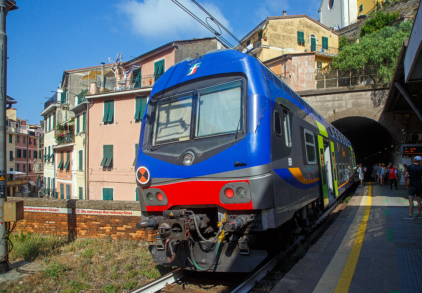 Ein Trenitalia Doppelstockzug (gezogen von einer E.464) hat am 22.07.2022, als Cinque Terre Express (Regionale) von La Spezia, via Riomaggiore, Manarola, Corniglia, Vernazza, Monterosso, nach Levanto, den Cinque Terre Bahnhof Vernazza erreicht. 

Vernazza ist eines der fünf Dörfer der Cinque Terre (Fünf Ortschaften) und verfügt, wie die anderen Dörfer, über einen Bahnhof an der Bahnstrecke Pisa–Genua (RFI Strecke-Nr. 77 / KBS 31 La Spezia–Genua). Der Bahnhof liegt zwischen zwei zweigeteilten Tunneln, jeder Bahnsteig hat seine eigenen Tunnelröhren. Wie auch in Riomaggiore passen die Bahnsteige nicht komplett unter freien Himmel, der Rest der Bahnsteige ist jeweils in einem der Tunneln. 