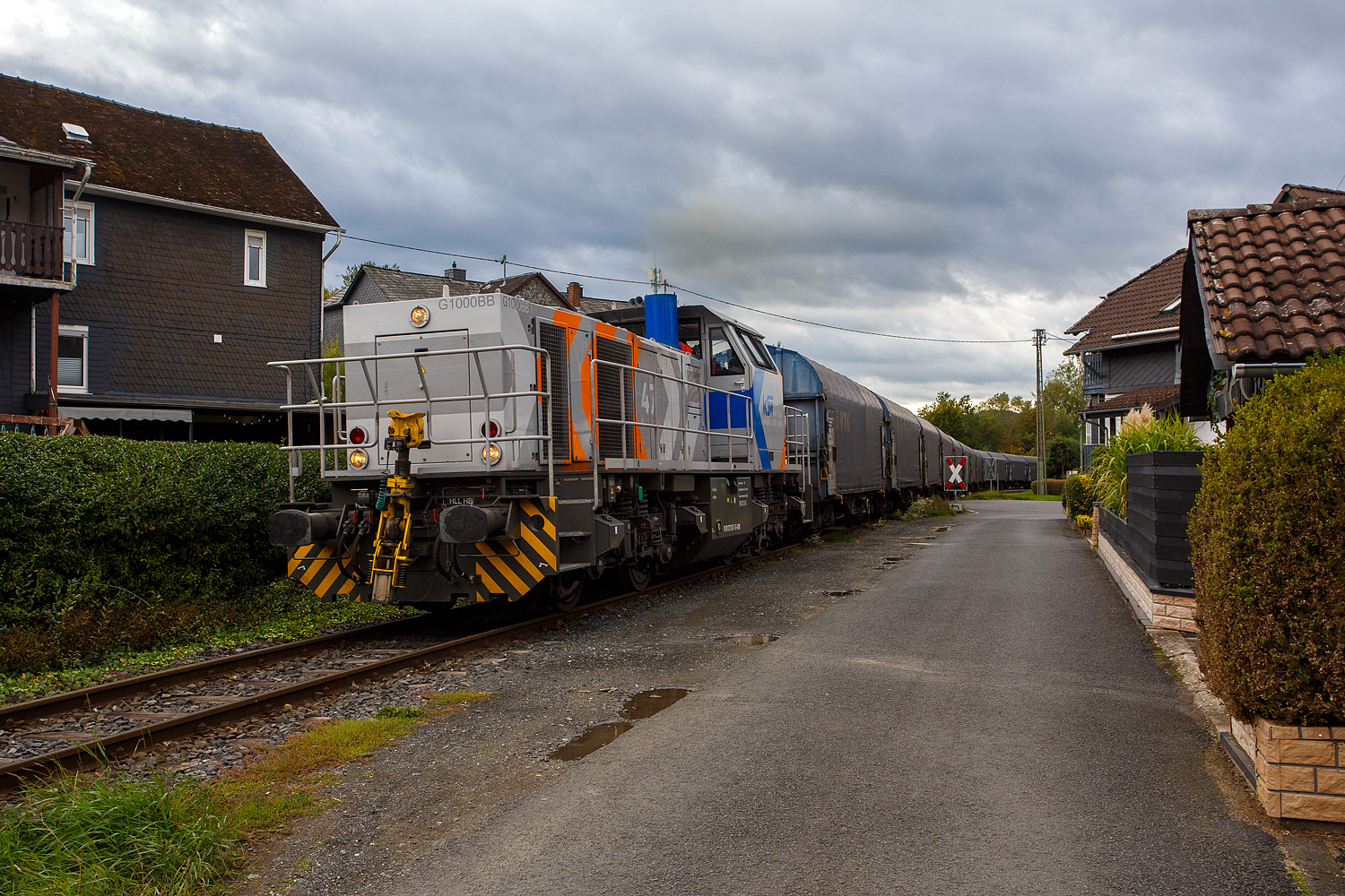 Nun hat die KSW 47 (92 80 1271 027-5 D-KSW), ex D 2 der HFM, eine Vossloh G 1000 BB der KSW (Kreisbahn Siegen-Wittgenstein), den Coilzug übernommen und zieht diesen nun vom KSW Rbf Herdorf weiter zu den Schäfer Werke KG am Pfannenberg in Neunkirchen-Salchendorf. Hier geht es gerade in Neunkirchen-Struthütten am 18 September 2025 zwischen den Häuser und Hecken hindurch. 

Auch wenn hierdurch, mehrmals täglich, die Anwohner genervt werden, zu Einem durch die Rollgeräusche und vor allem durch das erforderliche Pfeifen (Hupen) der Loks, wegen der vielen Bahnübergänge. Aber dennoch ist es schon lange praktifizierter Umweltschutz, denn mit allein dieser Zug ersetzt 40 bis 50 LKW. Übrigens diese Bahnstrecke der ehemaligen Freien Grunder Eisenbahn AG (auch bekannt als „Talbahn“) gibt es seit 1904, heute gehört sie als Betriebsstätte FGE -Freien Grunder Eisenbahn zur KSW Kreisbahn Siegen-Wittgenstein GmbH und wird als Freien Grunder Eisenbahn NE 447 (DB - Streckennummer 9275) geführt.

Mit einer eigenen Schieneninfrastruktur im Kreis Siegen-Wittgenstein, die mehr als 30 Kilometer umfasst, verfügt die KSW über wichtige Kapazitäten im wachsenden Güterverkehr. Auch wenn in Deutschland der Schienen-Güterverkehr geringer geworden ist, so ist er hier auf der „Talbahn“ die letzten Jahre ungemein gewachsen. 

Früher gab es ich Siegen (Eintracht) noch einen Abschnitt, wo die KSW bzw. frühere Siegener Kreisbahn auf der Eiserfelder Straße mit Läute (Glocke) fuhr, so wurde diese Bahn auch gerne im Volksmund „Bimmelbahn“ genannt.