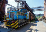 Die Dieselhydraulische Normalspur-Lokomotive Nr. 34, eine Henschel DHG 240 B  Esslinger , der Völklinger Hütte (ehem. Röchling'schen Eisen- und Stahlwerke GmbH in Völklingen/Saar, ab 1982 ARBED-Saarstahl GmbH, ab 1986 Saarstahl Völklingen GmbH) hier am 01 Juli 2025 abgestellt als Denkmal, mit zwei selbstentladende Schüttgutwagen der Gattung Fad oder Falns am Eingang der Rohstoffentladebunker in Richtung Hüttenbahnhof. Die Darstellung steht für den Rohstofftransport der Hütte zur Herstellung des Sintergutes. Die Lok war auch auf Bundesbahn Gleisen zugelassen. Heutiger Eigentümer der Lok und Wagen ist die Initiative Völklinger Hütte e.V..

Die zweiachsige dieselhydraulische Lok mit Antrieb über Gelenkwellen wurde 1969 von Henschel in Kassel unter der Fabriknummer 31210 gebaut und an die Röchling'sche Eisen- und Stahlwerke GmbH in Völklingen als Lok 34 geliefert. Weiterer Lebenslauf/Umfirmierungen: 
01.01.1971 > Stahlwerke Röchling-Burbach GmbH, Völklingen  34  
Juni 1982 	 > ARBED-Saarstahl GmbH, Völklingen  34  
Juni 1986 	 > Saarstahl Völklingen GmbH, Völklingen  34  
10.05.1989 > Saarstahl AG, Völklingen  34  
Später an MGS - Metallurgische Gesellschaft Saar mbH, Völklingen  34  
März 1999 	an Weltkulturerbe Völklinger Hütte - Europäisches Zentrum für Kunst und Industriekultur GmbH, Völklingen und Aufstellung als Denkmal. 

Die Lokomotive war bis zu einem Motorschaden 1997 in verschiedenen Bereichen eingesetzt: Transport von Duplexeisen zwischen der Mischerhalle und dem Siemens-Martin-Werk, im Bereich des Siemens-Martin-Werks, der Elektroöfen und des Hammerwerks sowie nach Einbau einer Funkfernsteuerung im Hafenbereich der Saarstahl AG.

Mit den Fahrzeugen der Typenreihe DHG 160 B / DHG 200 B / DHG 240 B und DHG 350 B bot Henschel Anfang der 1960er Jahre Gelenkwellenmaschinen für den unteren Leistungsbereich an, deren konstruktiver Ursprung nicht in Kassel, sondern in Esslingen der lag. Im Jahr 1961 übernahm Henschel das Diesellok-Typenprogramm der Maschinenfabrik Esslingen. Es wurde eine bei der Maschinenfabrik Esslingen als Typ Bern in Auftrag gegebene Lokomotive als DHG 325 in Kassel fertig gebaut und an den Auftraggeber, die Duisburger Kupferhütte, ausgeliefert. Der Typ Bern stellte auch die Basis für das oben genannte Typenprogramm dar. Dieses unterschied sich zwar optisch deutlich von seinem Esslinger Ursprung, übernahm aber die wesentlichen konstruktiven Merkmale der Fahrzeuge aus Esslinger Produktion. Gleichzeitig gab es keinen technischen und optischen Bezug zu den bisher im eigenen Henschel-Typenprogramm angebotenen Fahrzeugen.

Bei den neuen Gelekwellenfahrzeugen gab es leistungsmäßig deutliche Überschneidungen mit dem kleinsten Typ der 3. Generation, der stangengetriebenen DH 240 B. Diese wurde dann in der 4. Generation durch die DHG 240 B ersetzt.

Die Henschel DHG 240 B wurde zwischen 1963 und 1971 in 34 Exemplaren gebaut. Die Loks gingen vornehmlich an Chemiebetriebe und Raffinerien in Deutschland und der Schweiz. Von diesen wurden elf Loks explosionsgeschützt ausgerüstet. Einzelne Loks wurden ebenfalls mit Funkfernsteuerung ausgerüstet. Mit weitem Abstand am stückzahlenstärksten ist die DHG 240 B, die im Gegensatz zu ihren schwächeren  Schwestern  nicht mit einem DIWABUS-Getriebe von Henschel, sondern mit einem Voith-Hydraulikgetriebe ausgestattet ist. Zahlreiche Fahrzeuge stehen auch heute noch im täglichen Einsatz.

TECHNISCHE DATEN:
Baujahre: 1963 bis 1971
Gebaute Stückzahl: 34
Spurweite:	1.435 mm (Normalspur)
Achsfolge: B
Länge über Puffer: 7.160 mm
Achsabstand: 2.800 mm
größte Breite: 3.000 mm
größte Höhe über Schienenoberkante: 3.600 mm
Raddurchmesser: 850 mm (neu)
kleinster befahrbarer Gleisbogen: 35 m
Eigengewicht: 28 t
Max. Achslast: 16 t
Dieselmotor: wassergekühlter 6 Zylinder / 4-Takt-Diesel-Reihenmotor mit Turboaufladung, ohne Ladeluftkühlung vom Typ Henschel 6R 1416 A, Hubraum 13,3 l.
Leistung: 177 kW (240 PS)
Drehzahl: 1.600 U/min
Höchstgeschwindigkeit: 30 / 60 km/h
Leistungsübertragung: hydraulisch (Voit-Getriebe)
Kraftstoffvorrat: 400 l
